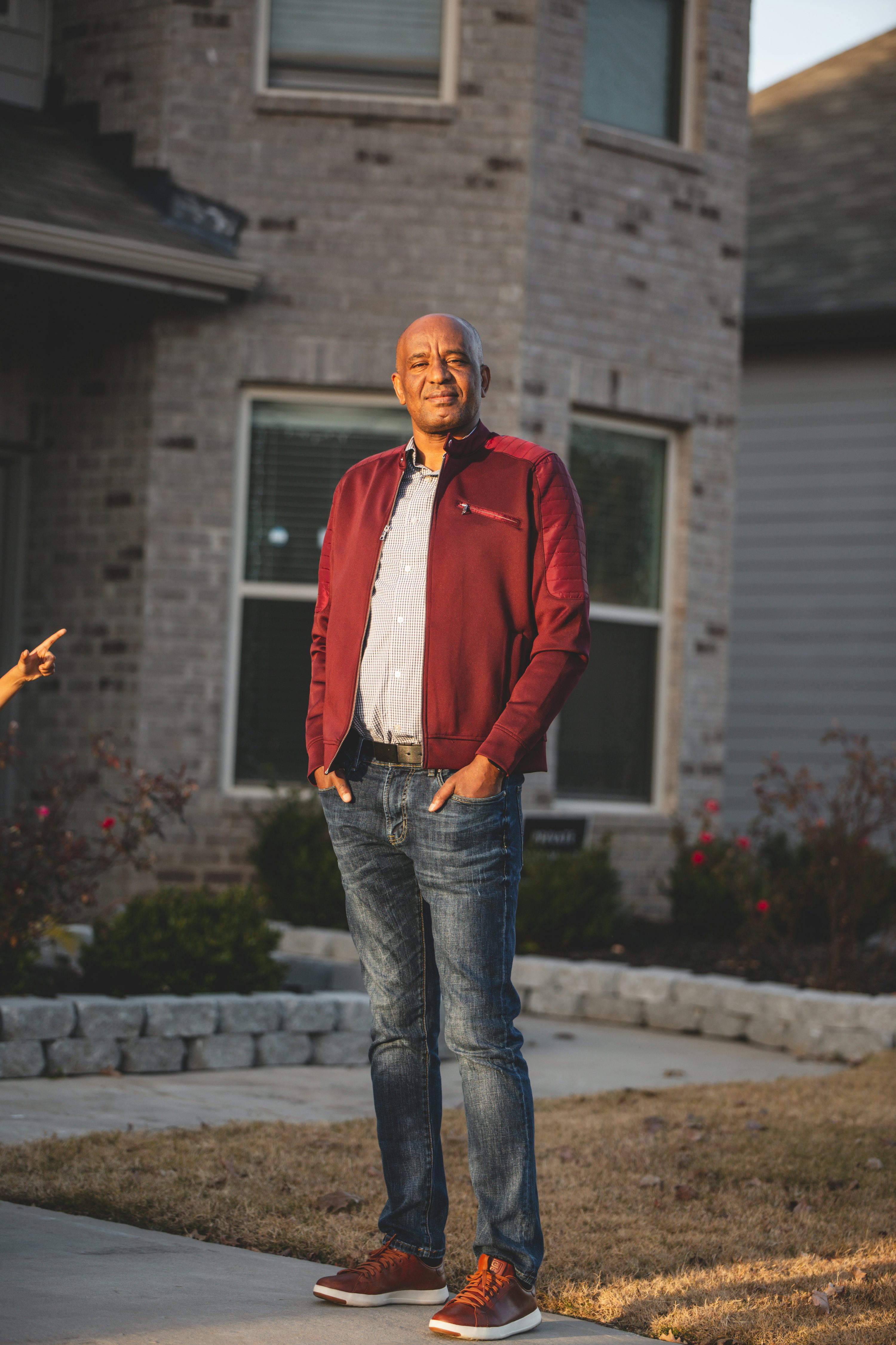 a smiling middleaged african american man stands in front of his house. he is wearing a red jacket, blue jeans, and brown shoes.  the sun is setting, casting a warm light on the scene.  the house is a modern, brick home with a wellkept lawn.  the man appears happy and content.