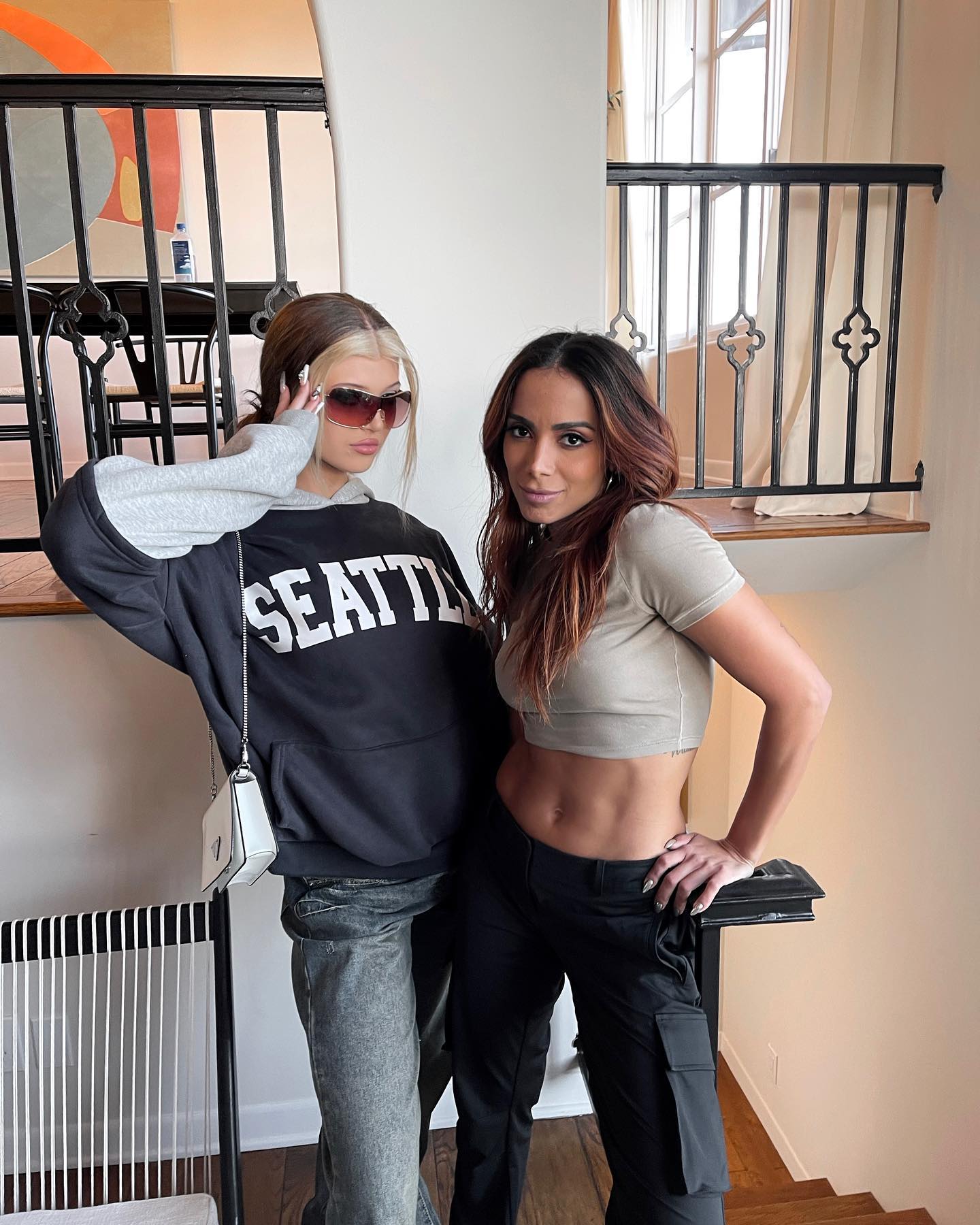 two women stand side by side indoors. on the left, loren gray wears a black hoodie with "seattle" printed on it, gray sleeves, light wash jeans, and sunglasses. she has a white purse on her shoulder. on the right, anitta wears a cropped beige tshirt and black cargo pants, with her hair styled in waves. they are standing near a staircase with a black railing.