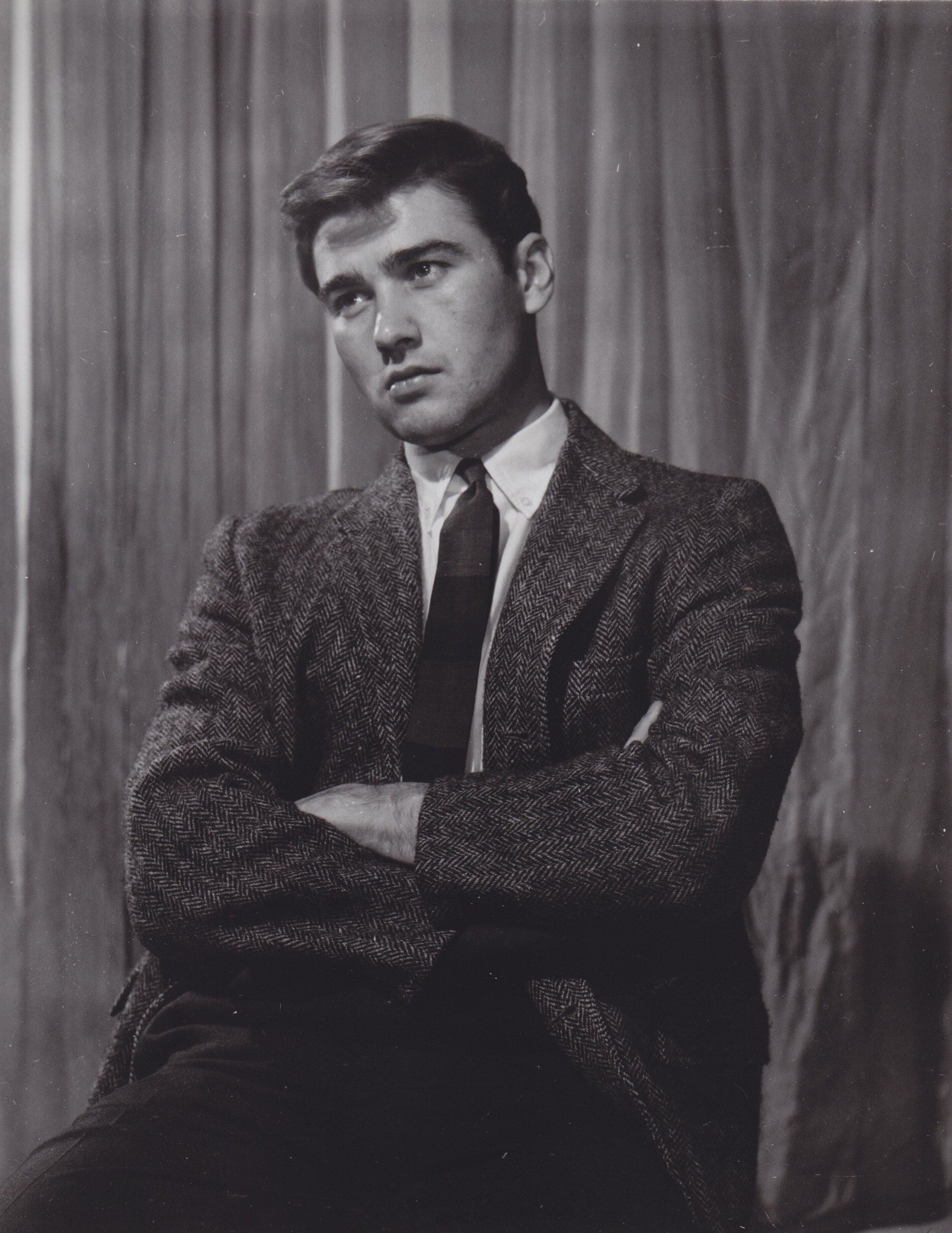 a black and white portrait of a young man in a studio setting. he is wearing a suit and tie, and his arms are crossed. the background is a neutral tone with vertical lines. he has dark hair and is looking up and slightly to the left.