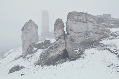 a grayscale image of a rocky landscape covered in snow, with a tower visible in the foggy background.