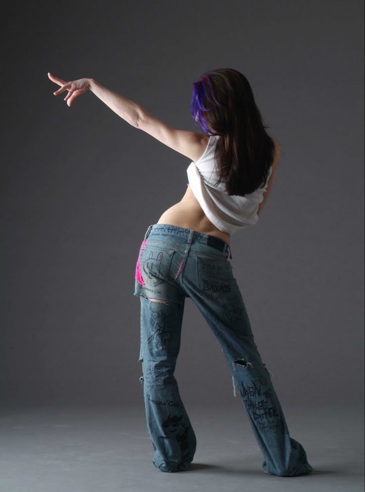 a full length studio shot of a woman dancing, viewed from the rear. she has dark hair with purple streaks and is wearing a white tank top and ripped jeans. her right arm is raised. she is barefoot.