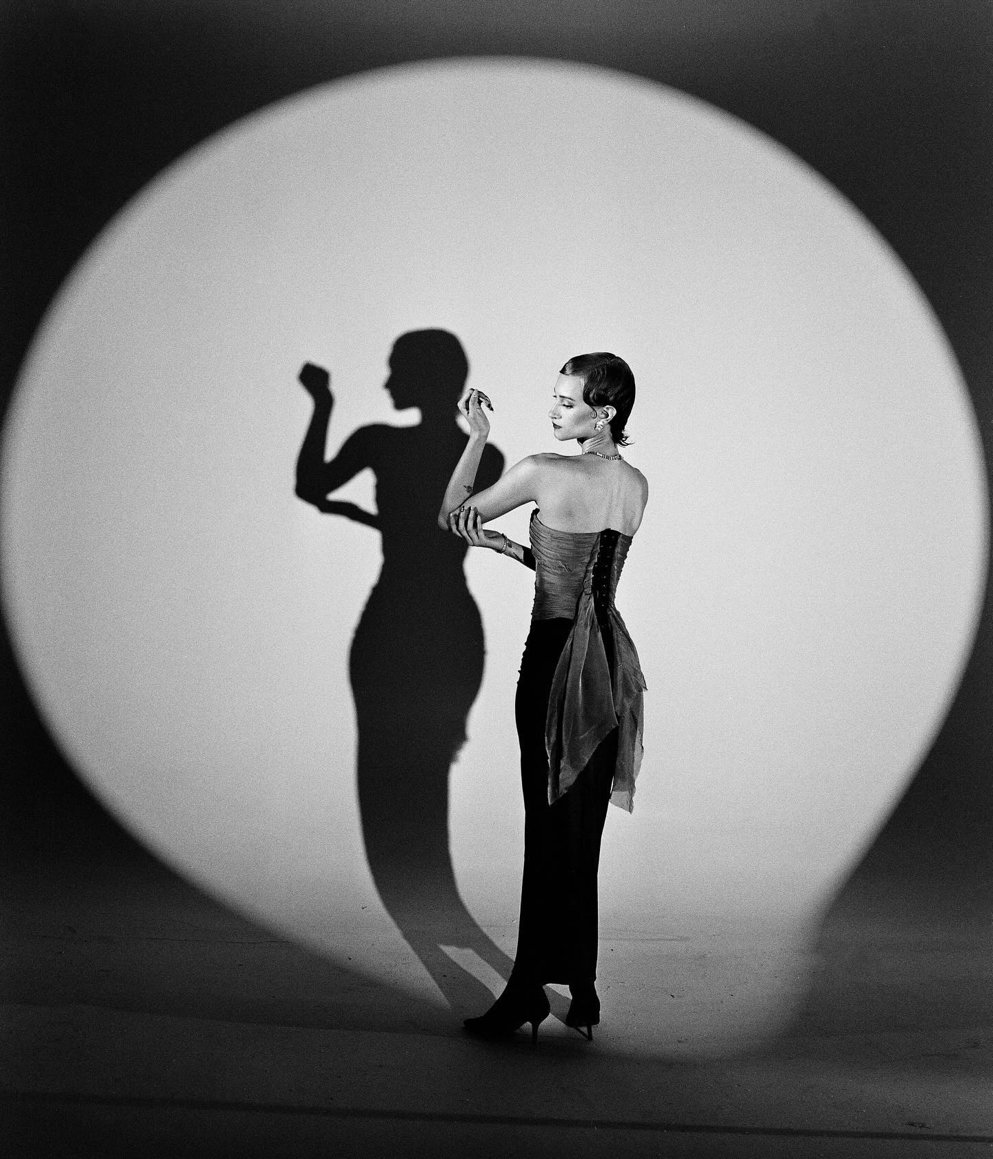 a woman in a glamorous black dress stands in a spotlight, her shadow stretching behind her. the image is in black and white and has a vintage feel.  the woman is posing, and the light creates a dramatic effect.