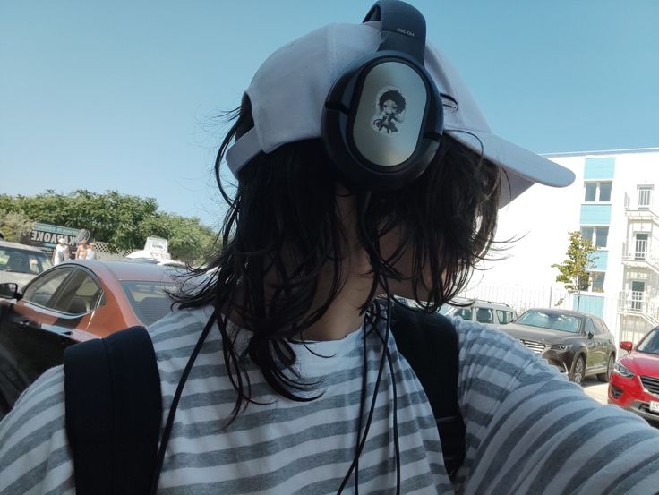 the image shows a closeup view of a person from the back, wearing a white hat, large overear headphones, a striped shirt and a backpack. their long dark hair flows down their back.  the person is standing on a street, and there are cars parked in the background. the setting seems to be an urban area on a sunny day. the overall style is casual.