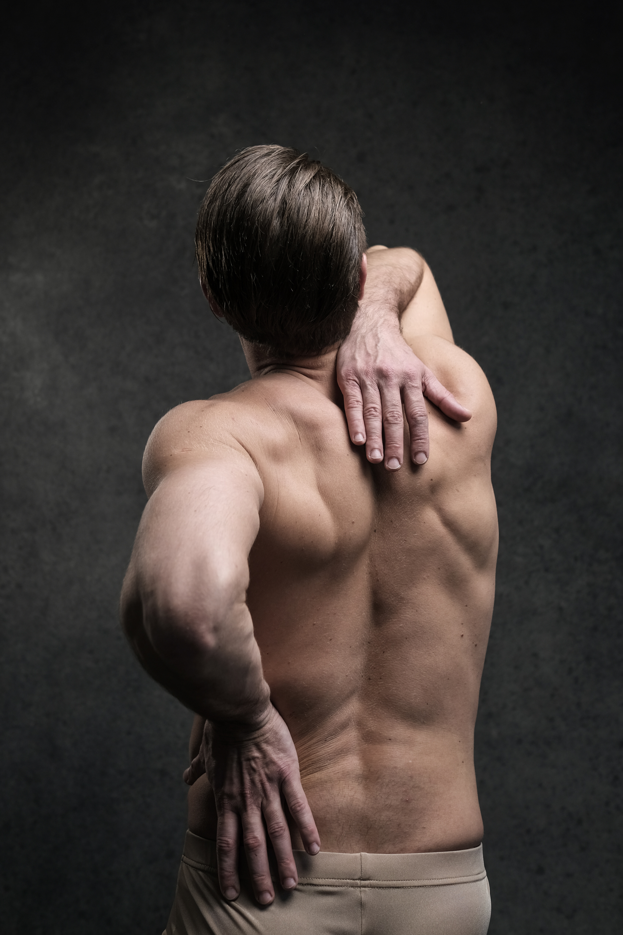 a shirtless, adult man is standing with his back to the camera, holding his lower back in pain. he has short brown hair and is wearing tan underwear. the background is a dark gray. the image suggests that the man is experiencing back pain.