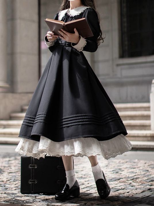 a person wearing a black lolita fashion dress is standing on a set of stone steps in front of a building. they are holding an open book and there is a suitcase next to them. the dress is black with a white lace petticoat, and the person is also wearing white socks and black shoes. they have long, dark hair and are wearing a white collar. the lighting is natural and the image is taken from eye level.