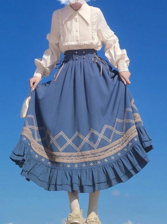 the image showcases a person, presumably female, from the knees down, wearing a stylish outfit against a bright blue sky. the focal point is the long, flowing blue skirt adorned with intricate embroidery forming a diamond pattern along the hemline and a delicate floral motif on the ruffled edge. the skirt is paired with a crisp, white blouse with puffed sleeves, tucked in to accentuate the waist. the low camera angle emphasizes the flow and volume of the skirt, creating a sense of movement. the bright, natural light enhances the vibrancy of the colors, particularly the blue of the skirt and the clear sky backdrop.