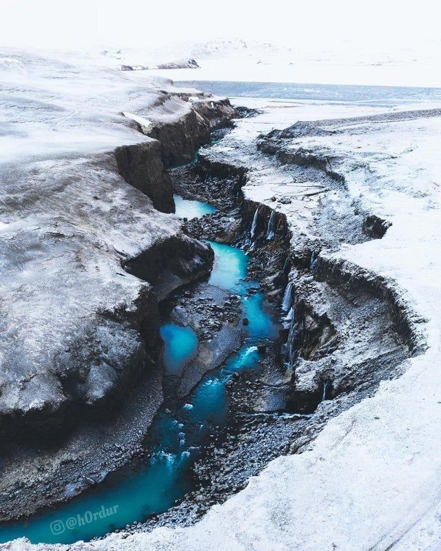 a highangle shot shows a river flowing through a snowy canyon. the river is a vibrant turquoise color, contrasting with the white snow and grey rocks. the canyon walls are steep and rugged, with patches of ice and snow clinging to them. the overall scene is cold and desolate, but the vibrant color of the river adds a touch of beauty to the stark landscape.