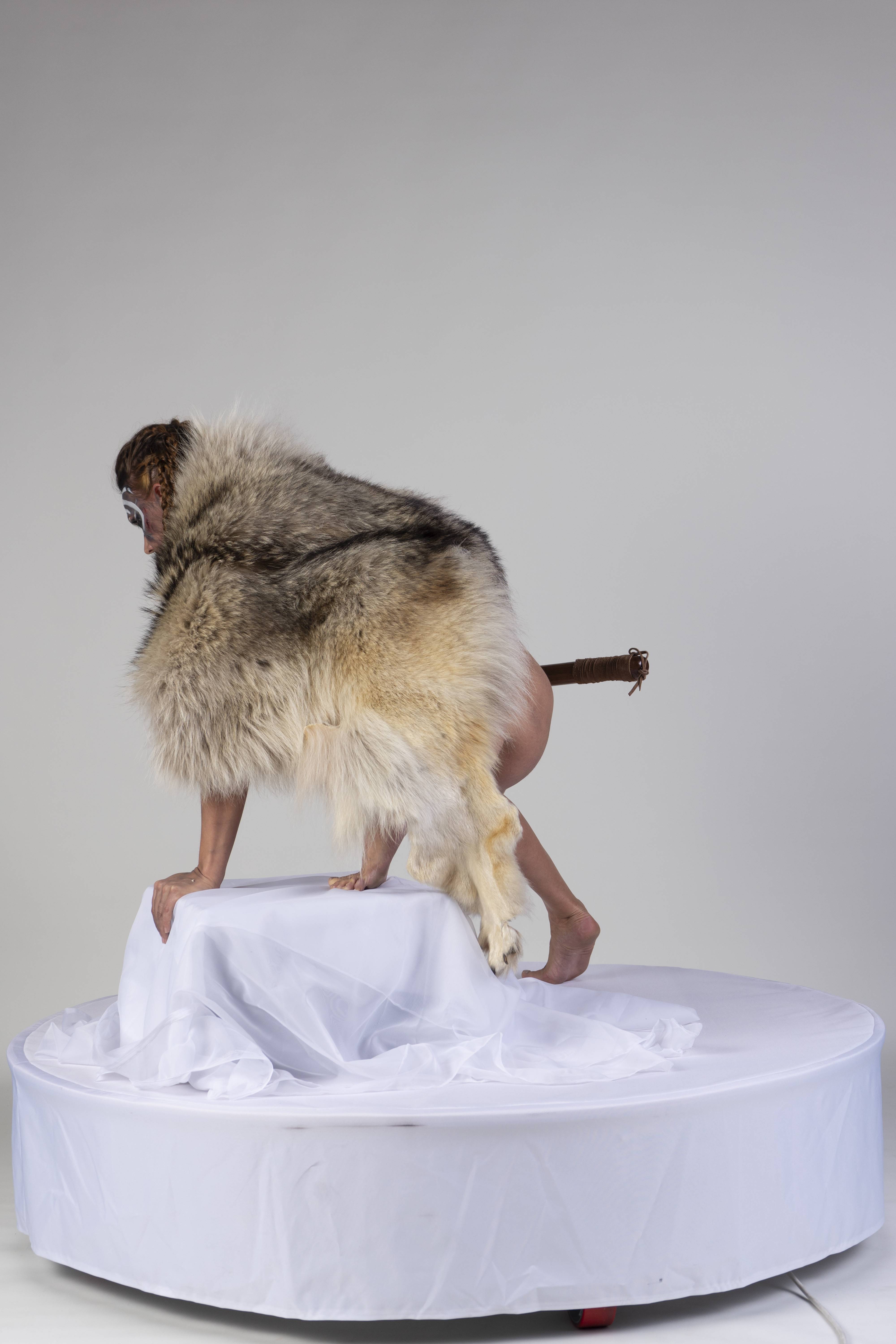 a person wearing fur and face paint is standing on a white platform in front of a white background. the person has a tail. the image appears to be a form of performance art. the person is in a crouched or kneeling position. the image could be considered bizarre, strange or unusual.