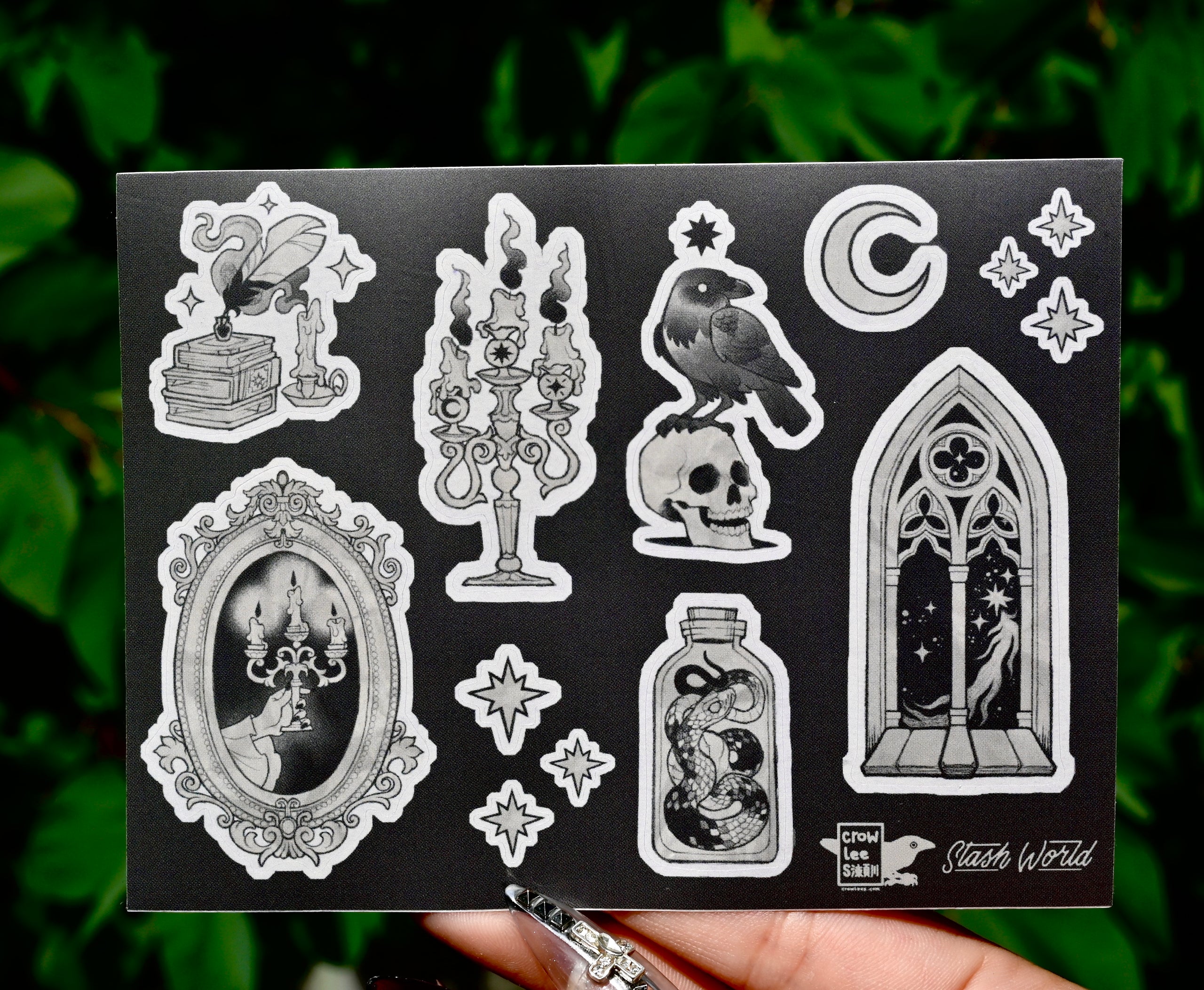 the image shows a closeup of a black sticker sheet featuring various gothic and occultthemed illustrations.  the stickers are in grayscale, and the designs include a raven perched on a skull, a candelabra with burning candles, a snake in a bottle, a quill and inkwell resting on books, a gothicstyle arched window with a celestial scene, a mirror with a candle holder, and several smaller details like stars and a crescent moon.  the sticker sheet itself is dark grey or black, providing a strong contrast to the light grey illustrations. the bottom right corner displays the artist's logo "slash world" and "crow lee studio". a hand is holding the sticker sheet in the bottom of the image, partially obscuring the bottom. the background is blurred, showing green foliage.
