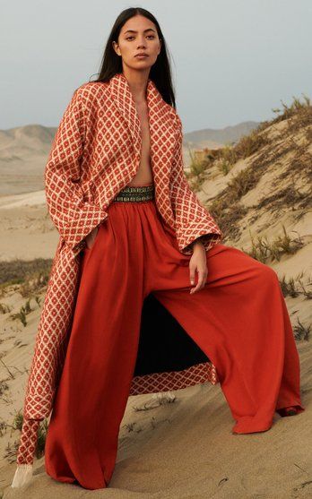 a fulllength shot of a woman wearing a stylish orange and offwhite patterned coat and matching wideleg pants. the coat has a geometric diamond pattern and an open front. the pants are a solid rich orange color. she is standing on a sandy beach or desert, with a serene background. the overall aesthetic is bohemian and elegant.  the model's long brunette hair is loose around her shoulders. she is posed in a relaxed but sophisticated manner.