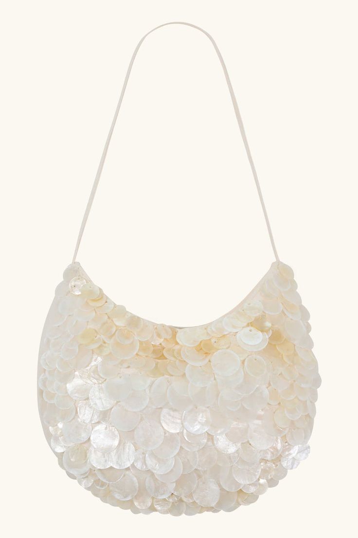 a closeup shot of a white sequin handbag with a strap against an offwhite background. the handbag is covered in round, iridescent white sequins that overlap each other. the strap is thin and white, and it extends from the top of the bag. the bag has a rounded shape. the background is a plain, light offwhite color.