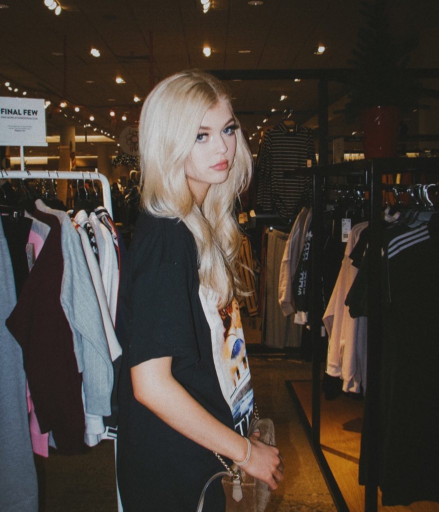 a young woman with blonde hair and blue eyes, identified as loren gray, is standing in a clothing store. she's wearing a black tshirt and carrying a purse. she is looking at the camera. clothes racks filled with merchandise are visible in the background, and there is a sign that reads "final few." the scene is lit with indoor lighting.
