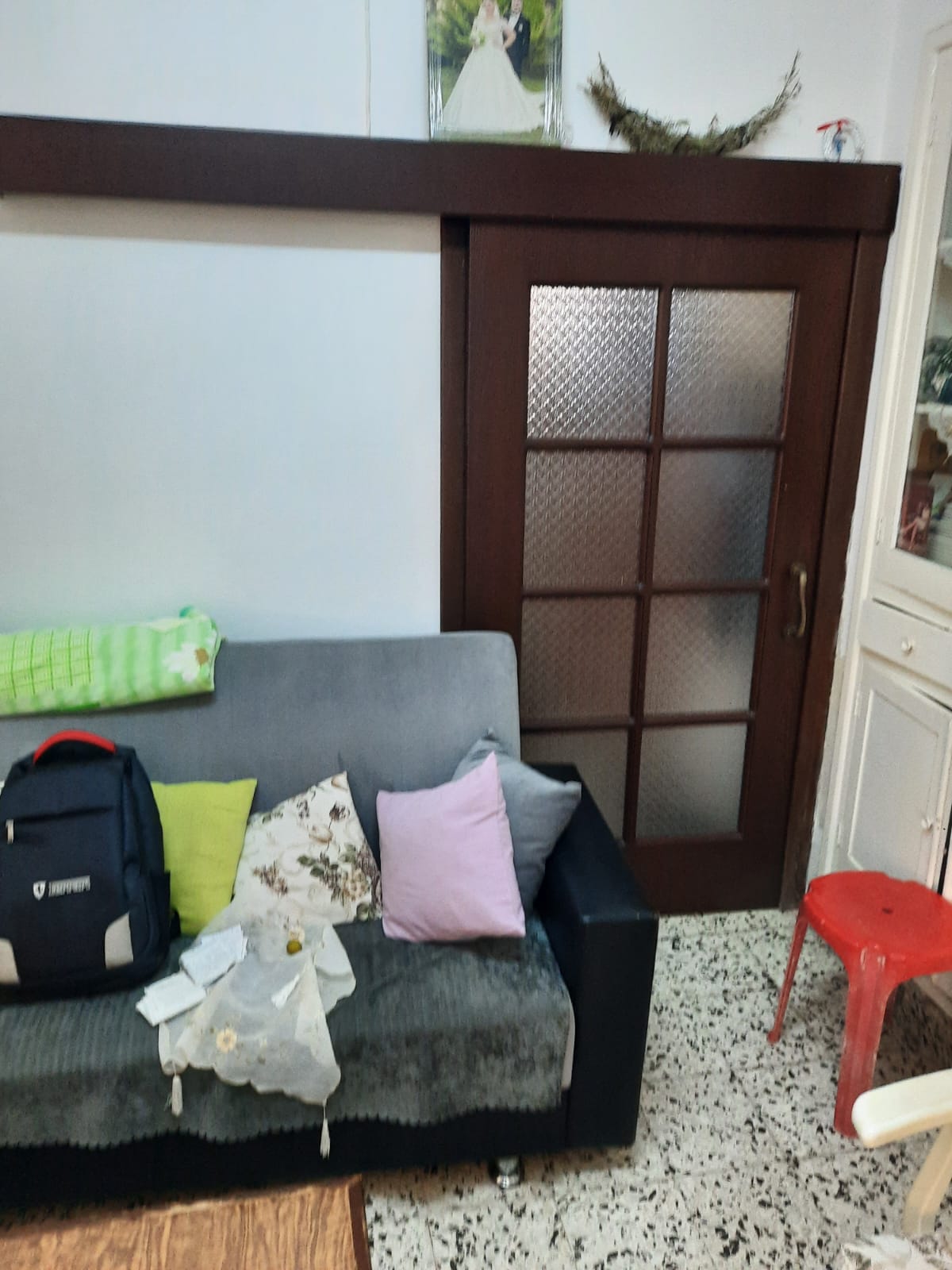 this image shows a room with a couch, pillows, a backpack, a door, a stool, and various home decor items. the couch is gray and has several pillows on it, including a pink one, a floral one, and a yellow one. a dark blue backpack is also resting on the couch. next to the couch is a brownframed glass door and a red plastic stool. above the door is a framed photograph and some other decorative items. the floor has a patterned design.