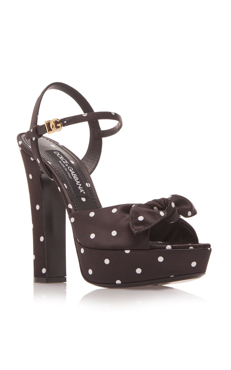 the image shows a single dolce & gabbana women's platform sandal.  the sandal is black satin with a white polka dot pattern. it features a peep toe, a platform sole, a high block heel, and a bow detail at the front. an ankle strap with a gold dolce & gabbana logo is visible. the shoe is positioned on a white background, with a subtle reflection visible beneath it.