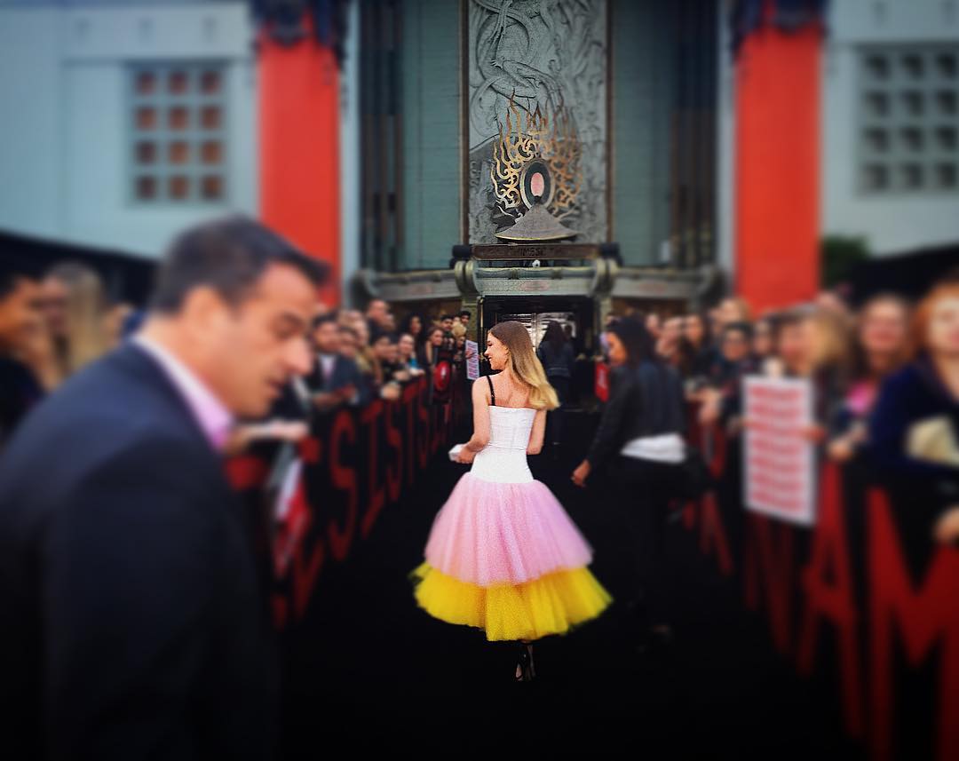 he image shows a woman in a pink, yellow, and white tulle dress at a red carpet event. she is standing with her back to the camera, and the background is blurry, showing a crowd of people and a building with red pillars. the overall mood is one of excitement and celebration.