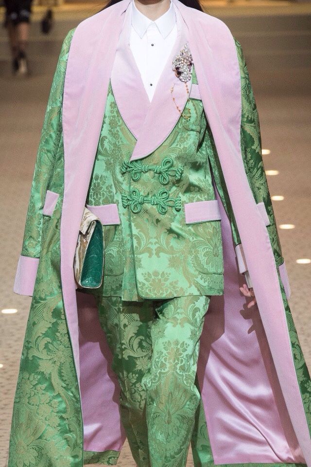 a runway model is wearing a green floral patterned suit with a pink lined coat, a brooch, and a green purse. the suit has a mandarin collar and a unique closure.