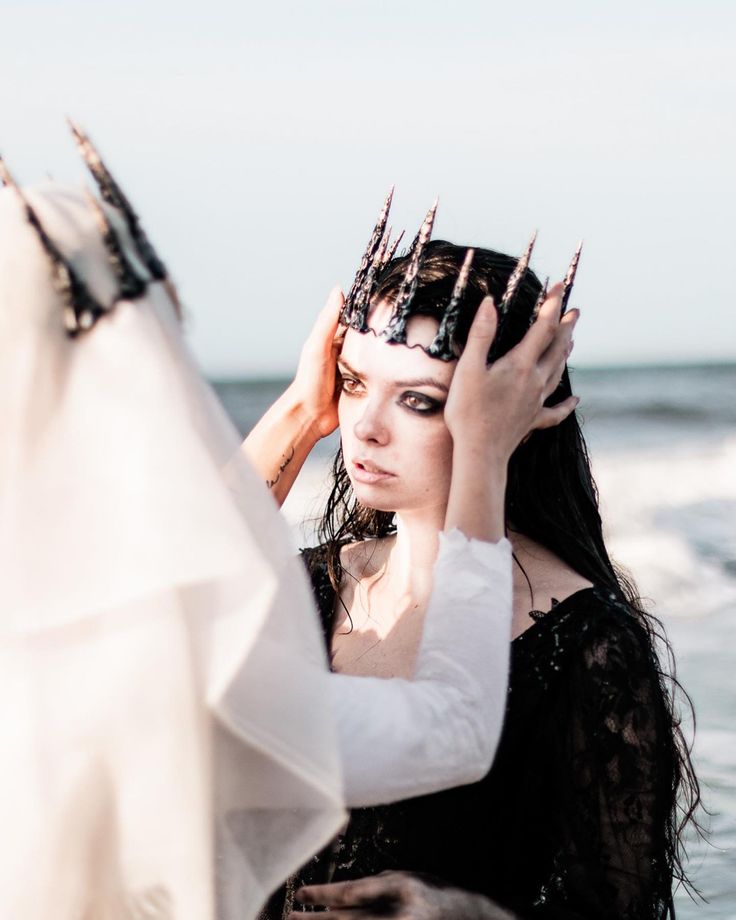 a woman with a crown on her head and a black dress stands on a beach looking away from the camera. she is being crowned by another woman in a white dress. the ocean is behind them. the image is taken in a warm, sunny day.