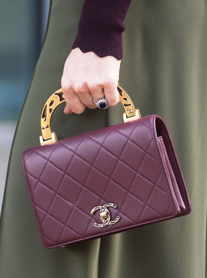 loseup image of a woman's hand holding a dark maroon chanel handbag. the bag features gold hardware, a unique top handle, and a quilted design. the woman is wearing a dark burgundy sweater and an olive green skirt. a sapphire ring is visible on her left ring finger. the focus is on the handbag and the woman's hand holding it. the image is welllit and sharply focused, showcasing the details of the bag and the woman's attire.