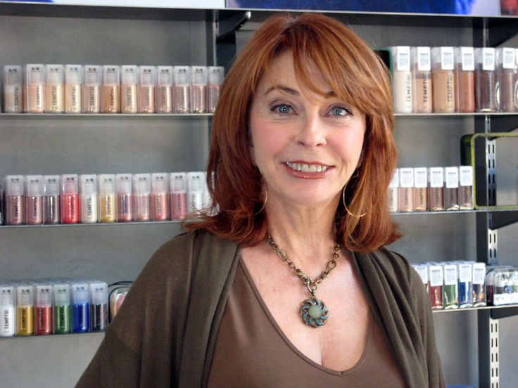a woman with long, red hair is standing in front of a shelf stocked with bottles of makeup. she is smiling at the camera and wearing a brown sweater and a silver necklace with a large green stone pendant. she has a natural, relaxed look and appears to be in a good mood.  she is likely a hairstylist or makeup artist working in a shop or store.