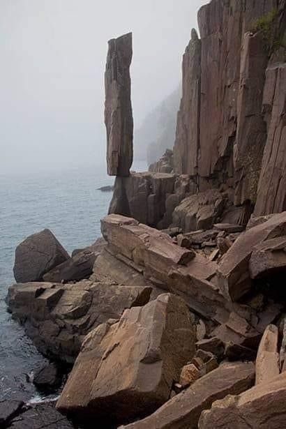 the image shows a tall, precariously balanced rock formation standing on the edge of a cliff along a rocky coastline. the ocean is visible in the background under a cloudy sky. the terrain is rugged and rocky, with various sized rocks scattered around. the landscape has a natural and weathered appearance.