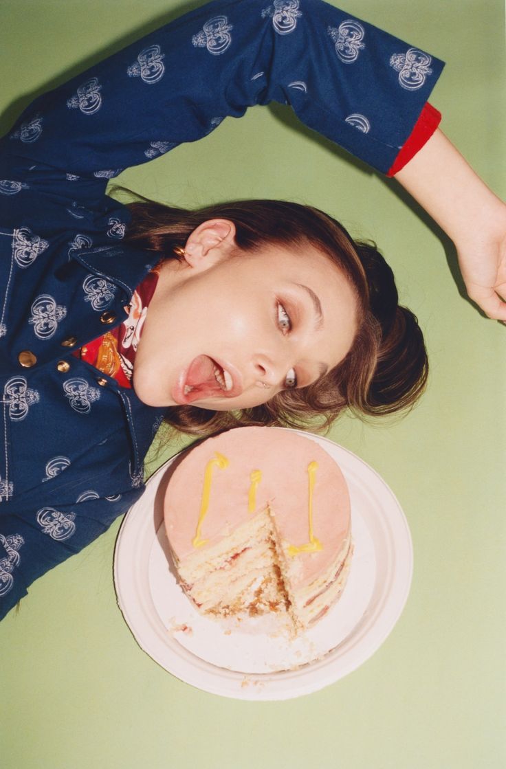 an overhead shot depicts a young woman lying on a pastel green background. she's wearing a blue patterned shirt with red cuffs and is looking towards a pink layered cake with yellow icing on a white plate.  her mouth is open in a surprised or playful expression. the overall style is trendy and fashionfocused.  the image appears to be a styled photoshoot, possibly for a fashion magazine or advertisement.