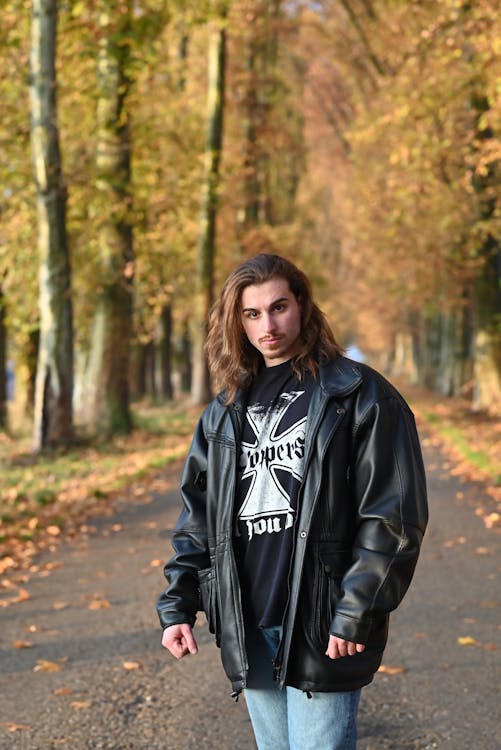 a man with long hair stands on a road lined with trees in autumn. he is wearing a black leather jacket over a black tshirt and blue jeans. the leaves on the trees are turning yellow and orange.