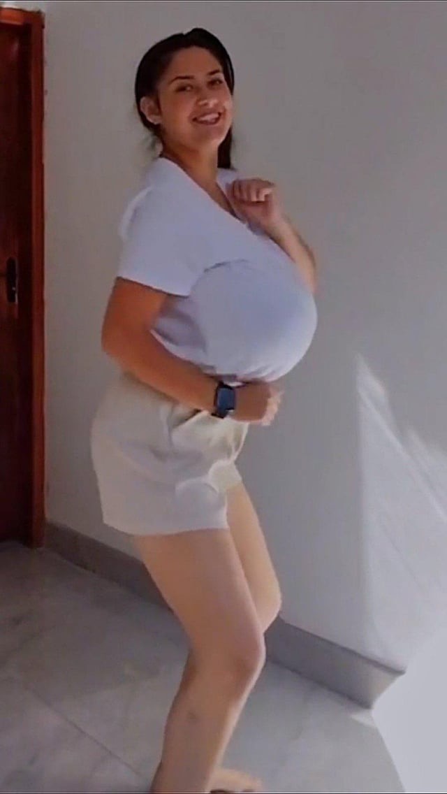 a young woman with brown hair is standing indoors and smiling for the camera. she is wearing a white shirt and white shorts. she is looking directly at the camera.