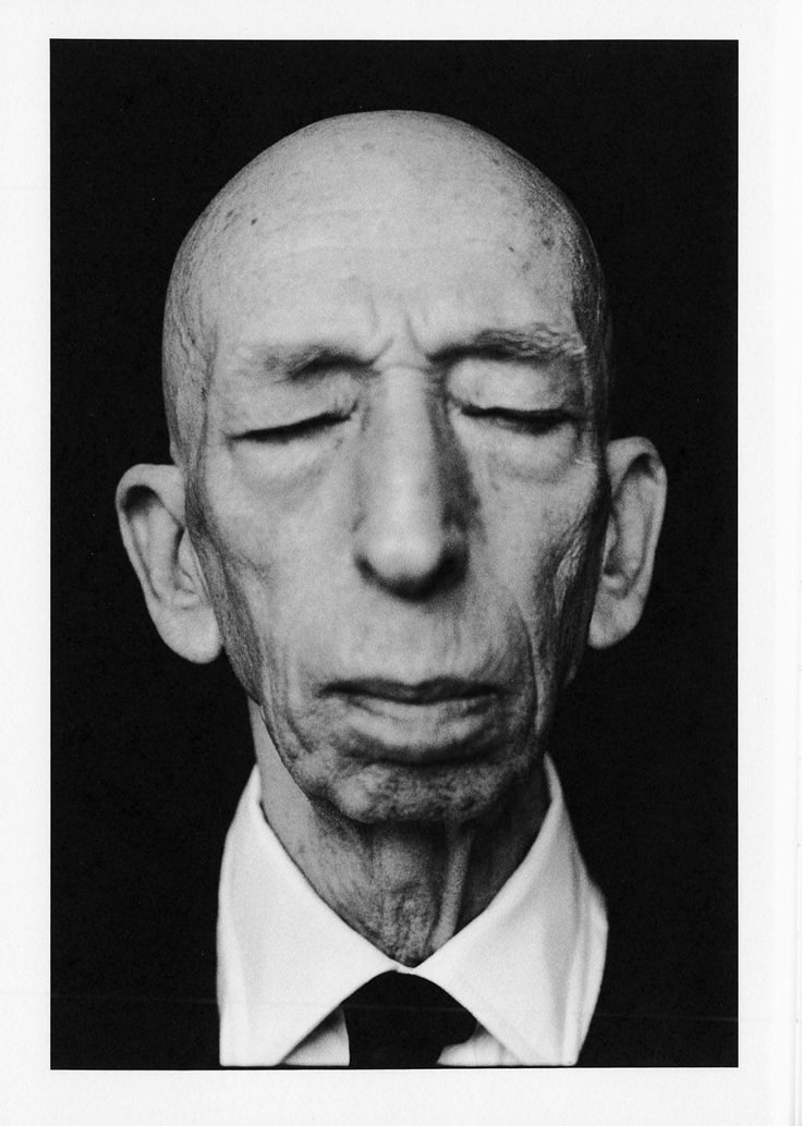 a black and white portrait of a bald, elderly man with his eyes closed, wearing a suit and tie. the image is a closeup, capturing the man's stern face and serious expression.