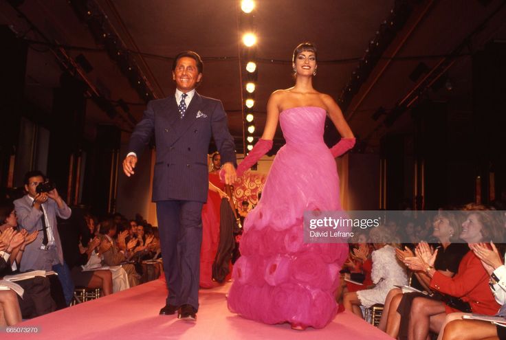 a fashion show features valentino and a model on the runway. valentino, dressed in a doublebreasted suit, walks alongside the model who wears a pink strapless evening gown and matching gloves. an audience applauds as they walk the catwalk. the event is welllit, showcasing the fashion designer and his couture creation.