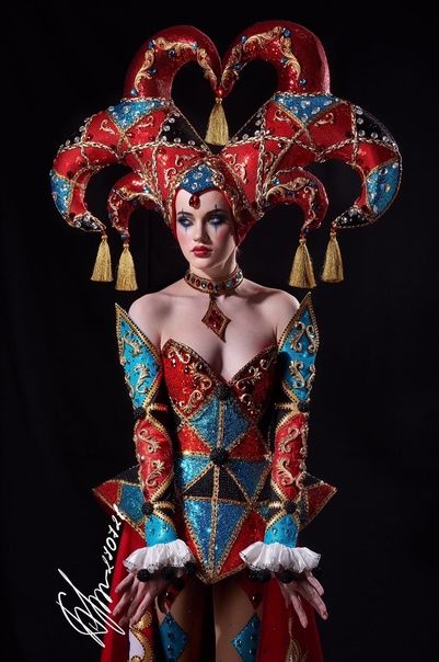 a woman dressed in a full jester costume stands before a black background.  her costume is very ornate, made of red, black, blue, and gold fabric, sequins, and diamond shaped patterns.  she wears a matching headpiece and has dark eye makeup.  the costume looks elegant and fancy, and is theatrical in nature.