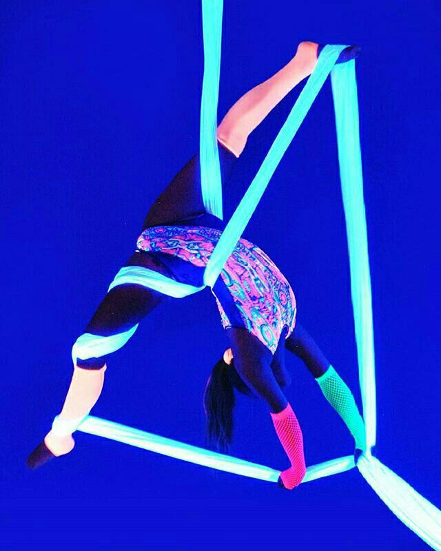 a woman is performing on an aerial silks apparatus. she is midair, in a pose that shows her strength and flexibility. the silks are neon blue and glow in the dark, creating a vibrant and eyecatching image. the background is a solid blue, which further emphasizes the vibrant colors of the silks and the woman's attire. the woman appears to be smiling, as if she is enjoying the performance.