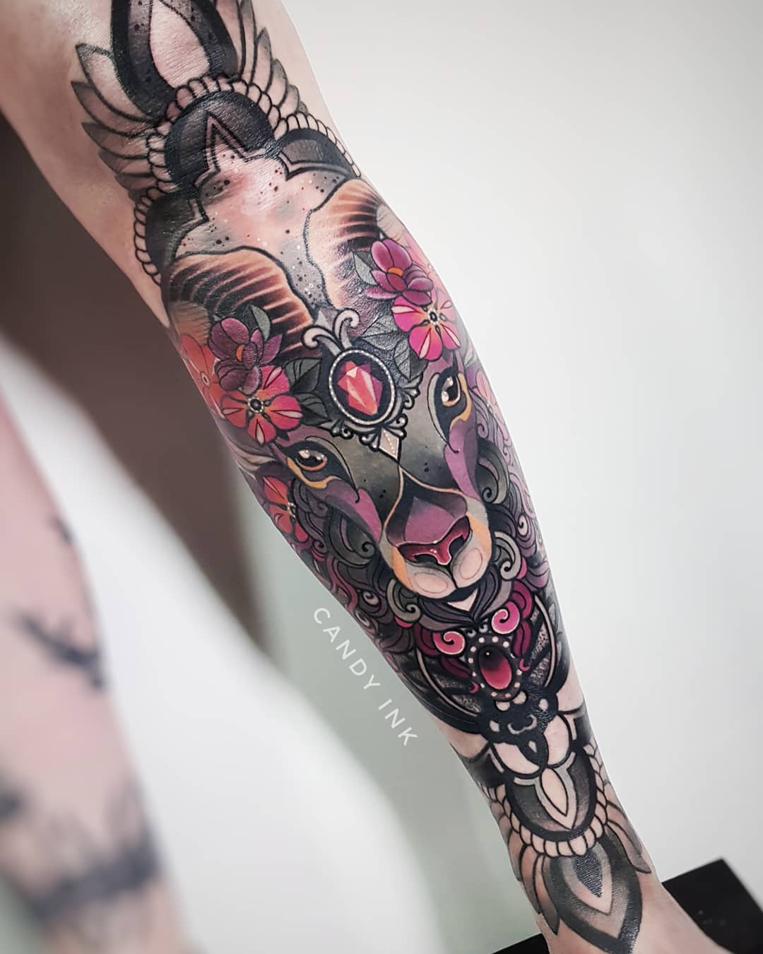 a closeup photograph showcasing a vibrant neotraditional tattoo of a ram's head adorned with flowers and jewels on a person's arm.