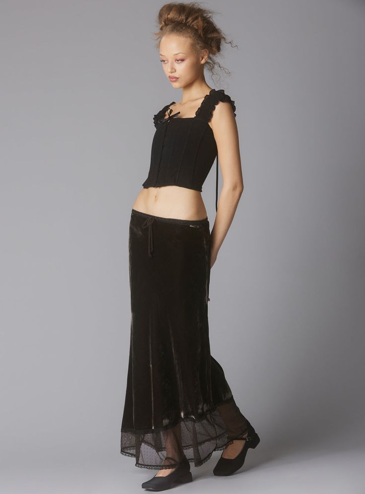a fulllength studio shot of a woman modeling a dark brown velvet maxi skirt paired with a black corset top. the skirt has a sheer, lacelike trim at the bottom. the corset has ruffled straps and a tie at the center. the woman is wearing dark flat shoes and has her hair styled in an updo. the background is a muted grey. the overall aesthetic is elegant and slightly gothic.