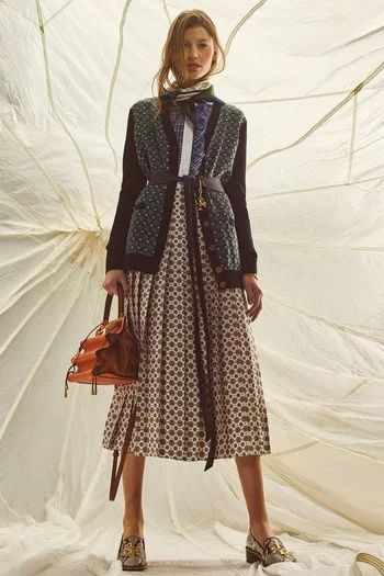 a full shot of a fashion model posing for a photoshoot. she is wearing a patterned pleated midi skirt, a dark green and black patterned cardigan, a brown belt, and loafers. she carries a small orange handbag and has a scarf wrapped around her neck. the background is a white fabric backdrop with folds and creases. the overall style is autumnal and sophisticated.