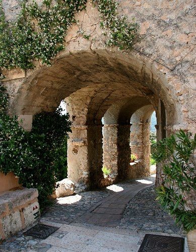 the image depicts a stone archway with multiple arched sections, leading into a village street. the archway and columns are made of rough stone, with plants and greenery growing on and around them. the street is paved with cobblestones and stone slabs. light and shadow play across the scene, highlighting the texture of the stone and foliage.