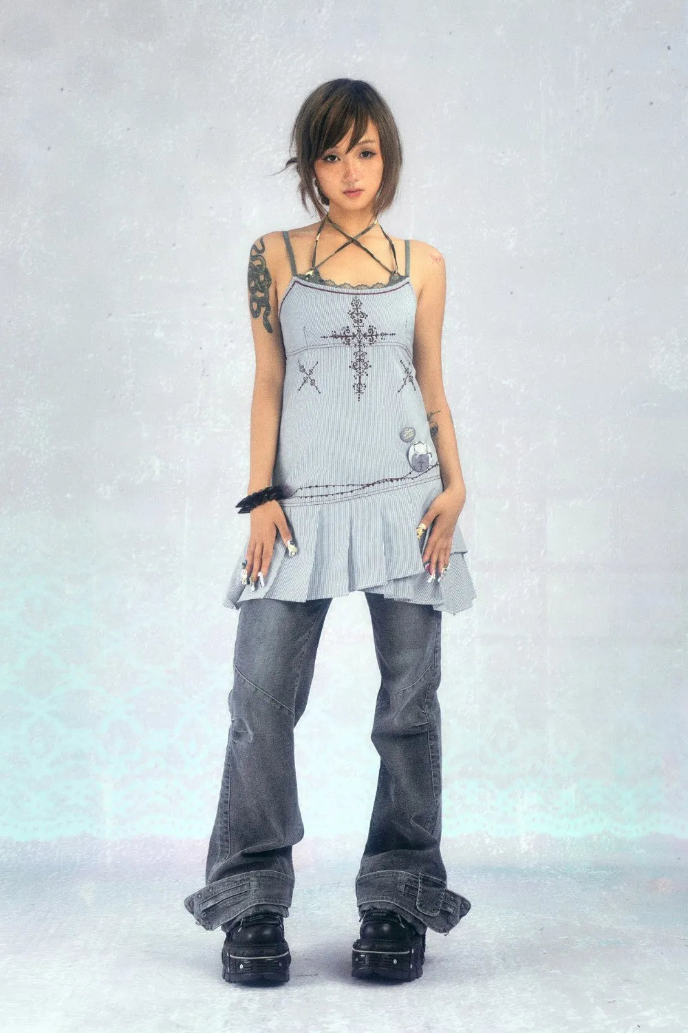 a full shot of a young japanese woman with a bob haircut wearing a light blue, embroidered dress over a pair of dark gray jeans and chunky dark boots.  she has visible tattoos on her arm and is posed in a studio setting against a light gray background.  her expression is neutral and her overall style is fashionable and somewhat edgy.