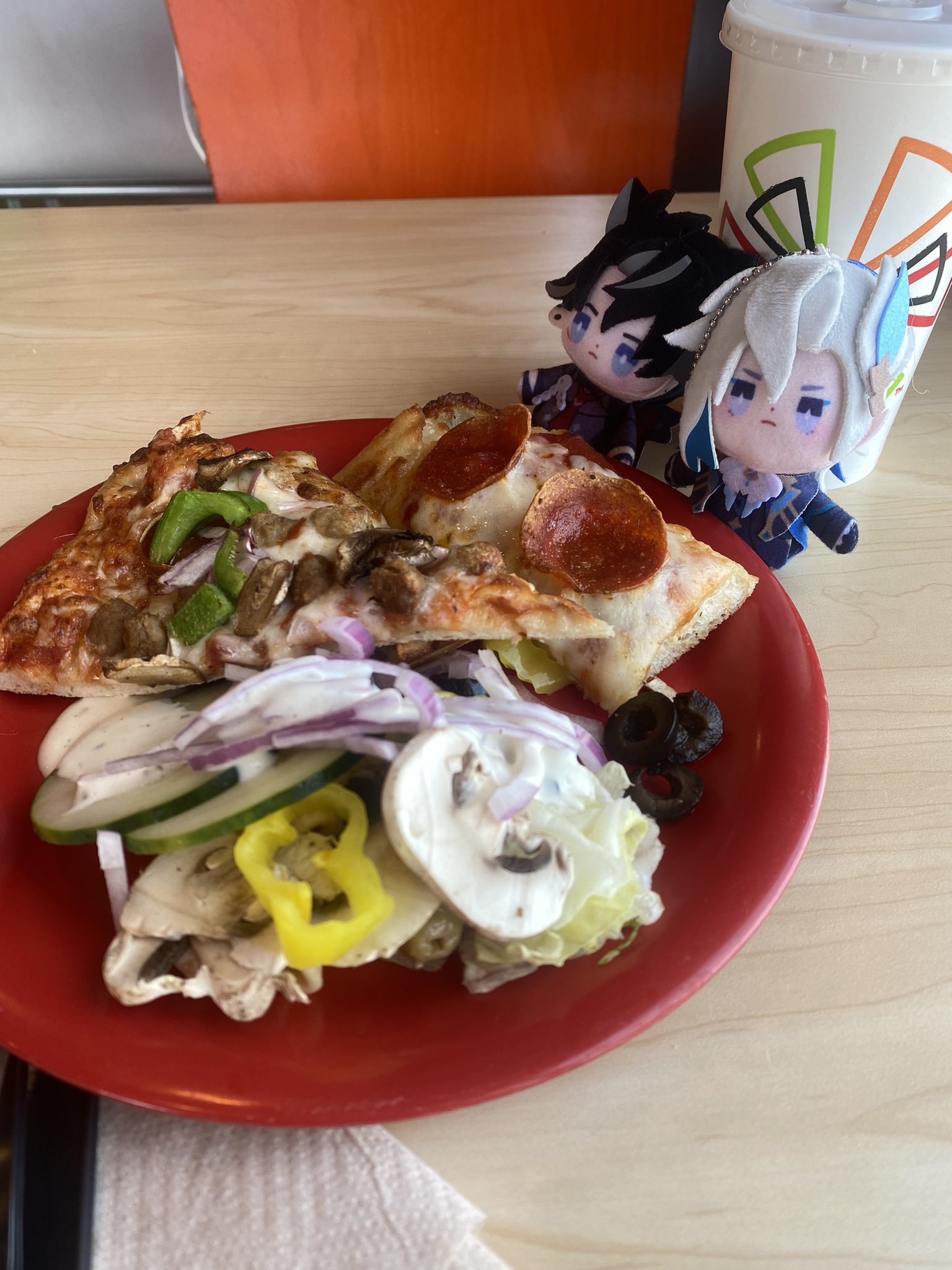 a closeup shot of a pizza with a side salad in a red plate, a couple of anime figurines are behind it. the pizza is cut into triangles and has pepperoni, mushrooms, and other toppings. the salad is made with cucumbers, red onion, peppers, mushrooms, and a creamy dressing. the pizza and salad are on a table in a restaurant.
