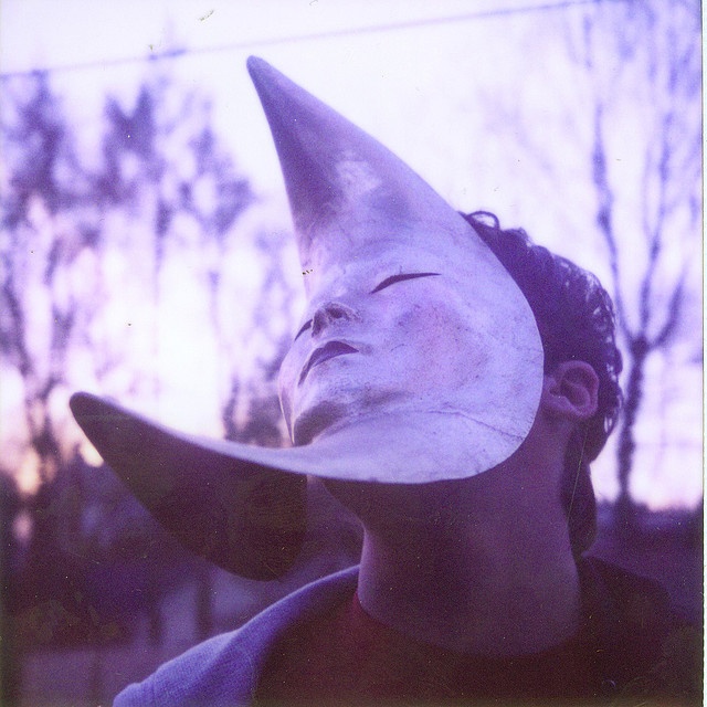 a young man is wearing a silver crescent moon mask. the mask covers his entire face and has his eyes closed. the man's hair is dark and pulled back from his forehead. the image is blurred and appears to be taken with a camera phone. the background is blurry and out of focus.  the overall color scheme is purple and silver. the image has an artistic quality to it.