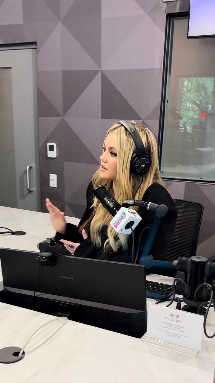 avril lavigne wearing headphones and speaking into a microphone in a radio station studio setting. she is seated behind a table with a samsung monitor and a guest wifi sign. the background features a patterned wall.