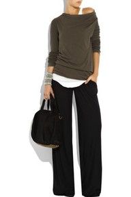 a woman is wearing a green top with a black, loosefitting pants. she is also carrying a black bag, with silver buckles. she is wearing bracelets on her wrists. she is staring at the camera and has a casual, elegant style.