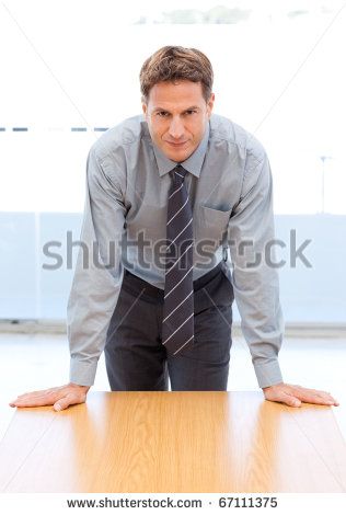 a man wearing a grey shirt and a dark tie looks at the camera with a serious expression. he is standing with his hands on a brown desk.  he is probably a businessman in a corporate setting.  he is confident and looks to be ready to lead.