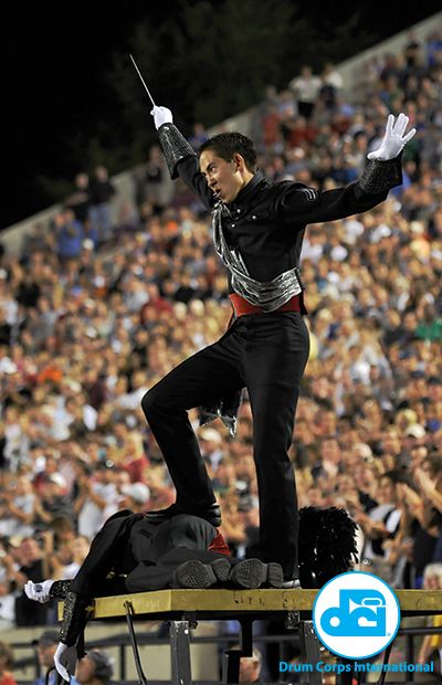 a drum corps conductor in a black uniform with a silver sash and white gloves is leading a marching band during a performance. the conductor is standing with one leg raised and is holding a baton in his right hand. the conductor is looking at the band members as they perform. the band is in front of a large crowd of people. the image is taken from a low angle, looking up at the conductor and the band. the image is brightly lit and has a lot of detail. the image is a good representation of the excitement and energy of a drum corps performance.