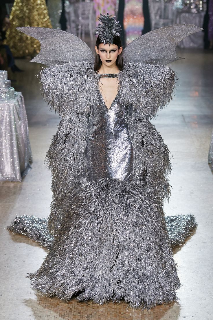 a model walks down a runway wearing a silver couture gown. the gown is elaborately decorated with tinsellike fringes. she also wears large silver wings and a matching headpiece. her makeup features dark eyeliner and black lipstick. the dress has a deep vneckline. the fashion show setting is visible in the background.