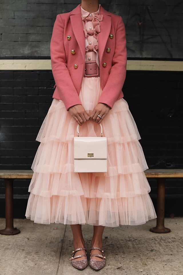 fulllength shot of a woman standing outdoors, showcasing a stylish outfit. she's wearing a blush pink doublebreasted blazer over a delicate, layered tulle skirt in a similar shade.  underneath the blazer, a blouse with a bow detail is visible. the skirt is a voluminous, multitiered tulle design. she accessorizes with a small, white rectangular handbag and sparkly rosegold mary jane shoes.  the woman’s style is elegant and feminine, creating a sophisticated and playful look. the background is a dark brick wall and a wooden bench.