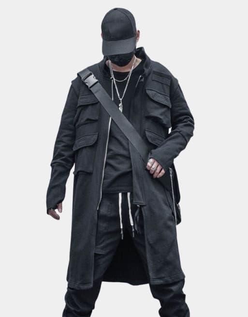a man dressed in an allblack stylish street outfit is standing in a studio setting. he is wearing a black jacket with multiple pockets, a black shirt underneath, black pants, a black cap, and sunglasses. he has a crossbody bag that has a black strap and a black bag.