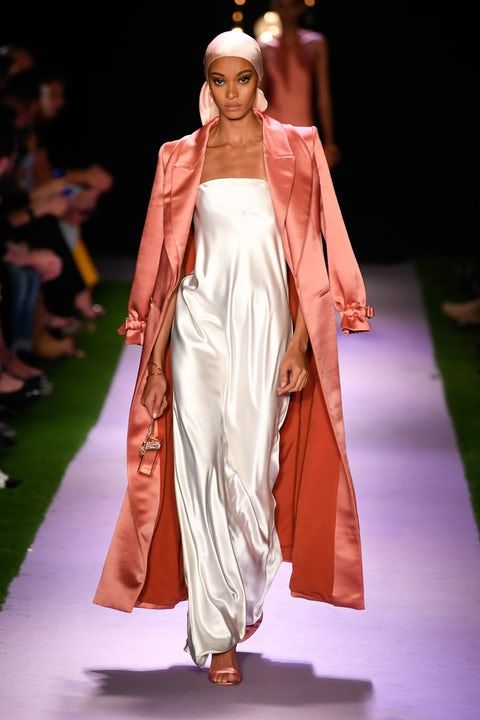 a model walks down a runway wearing a long, coralcolored satin coat over a white silk slip dress. she is also wearing a pink headscarf and delicate sandals. the runway is a lavender color, and the background is slightly blurred but shows an audience. the overall style is elegant and glamorous.
