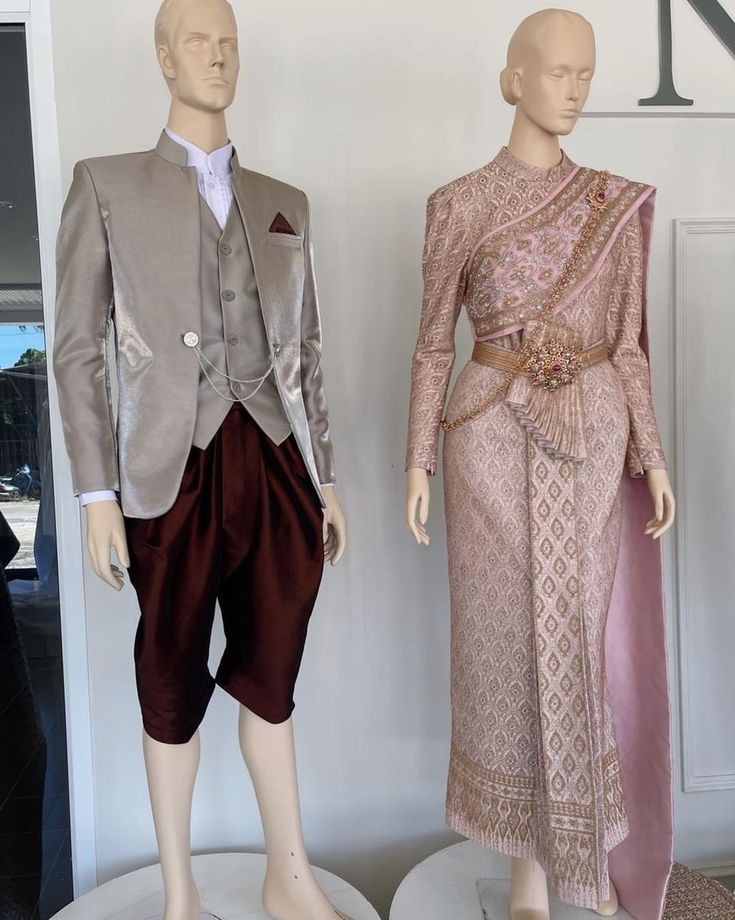 a thai bride and groom stand side by side, wearing traditional wedding attire. the bride is in a beautiful pink dress with intricate details, while the groom is wearing a  grey suit with maroon pants. they look elegant and happy as they prepare for their special day.  the image captures the beauty and cultural significance of thai weddings.