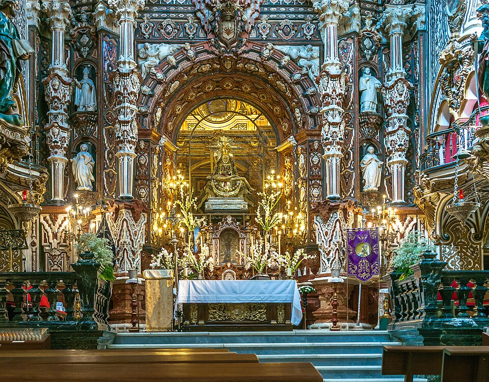 the image shows a highly ornate altar within a church. the architecture is predominantly baroque, featuring intricate carvings, sculptures, and details in gold, white, and dark wood tones. columns flank the central area, adorned with statues. candles illuminate the altar, which is covered with a white cloth and decorated with plants. a religious banner is also visible. the overall atmosphere is one of opulence and religious reverence.