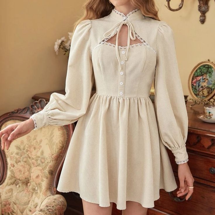 a woman is modeling a vintageinspired dress. the dress is a light beige corduroy with long sleeves, a high collar, and a tie at the neck. the dress also has a laceup detail on the bodice and a buttonfront closure. the woman is wearing her hair down and is standing in front of a mirror. the room she is in is decorated in a vintage style with ornate furniture and a floral rug.