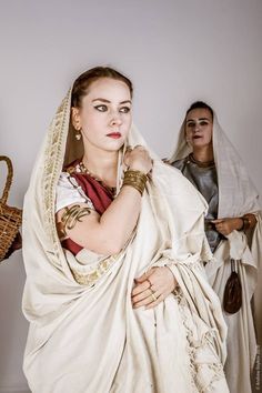 two women are standing indoors wearing roman style offwhite togas. the woman on the left is in the foreground looking left. the woman on the right is in the background looking directly at the camera.